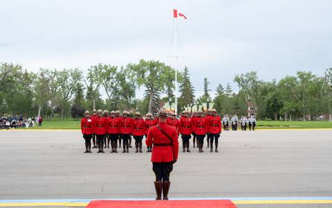 A troop of RCMP cadets in red serge stand together and look right. There is a drill instructor giving direction and there are members of the RCMP Cadet band and the public in the background.