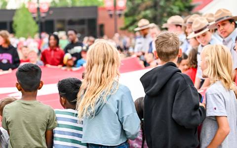 A large group of children along with RCMP cadets in Stetson hats unfurl a large Canada flag.