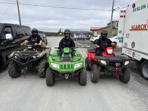 Two uniformed RCMP officers and one uniformed Conservation Enforcement Officer are geared up and heading out on ATV patrol. Marked police vehicles and a search and rescue command post vehicle are seen in the background.