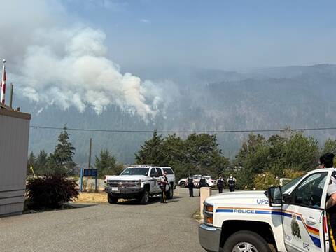 RCMP vehicles and officers near a road as heavy smoke from a wildfire rises from a forested mountainside in the background.