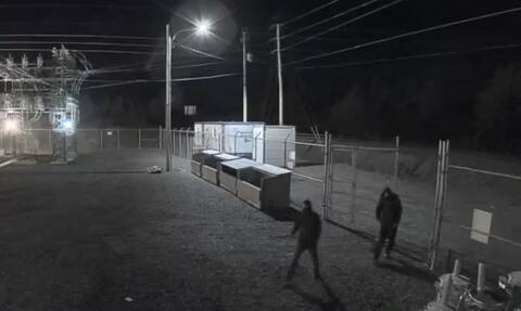 Two individuals, dressed all in dark clothing with hoods over their heads, walk into a fenced compound at night. A power substation is visible in the background.