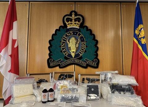A table placed in front of an RCMP crest on the wall shows items seized drug a search of a suspect’s home. The drugs are displayed in see-through plastic bags and there are three black handguns also shown.