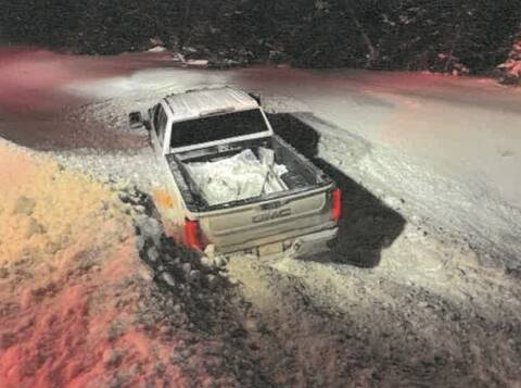 a light-colored GMC pickup truck is stuck in deep snow, off the side of a snow covered roadway. Trees are visible in the background.