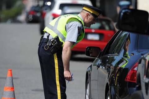 RCMP police officer bent over at the hip, talking with the driver of a black vehicle through driver side window during a road check