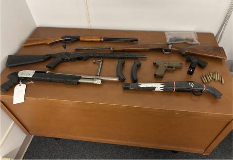 Items seized by police laying on a table, including three rifles, a sawed-off shotgun and ammunition. // Les objets saisis par la police sont posés sur une table, notamment trois fusils, un fusil à canon scié et des munitions.
