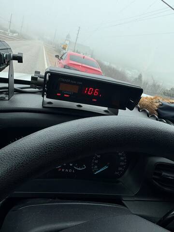 A red-coloured car stopped on the side of the road. In the foreground, a police radar shows a speed reading of 106 km/hr.