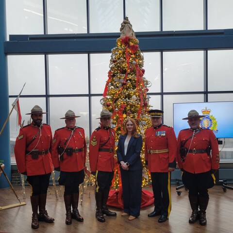 Five RCMP officers pose with a woman from the Make a Wish foundation in front of a large tree.