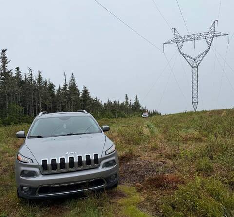 A grey Jeep SUV sits below transmission lines, with a marked police vehicle visible in the distance.