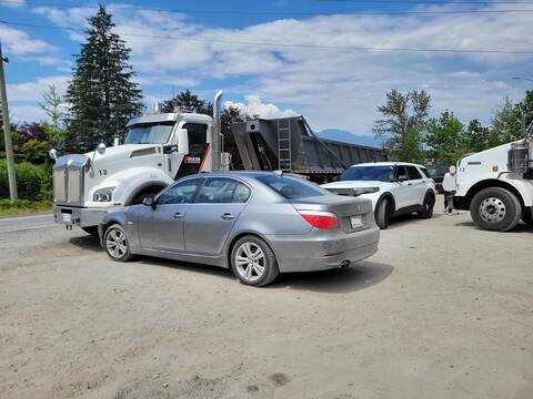 A grey BMW sedan sits entangled with a white dump truck in a dirt parking lot. A BC Highway Patrol unmarked police vehicle is in the background