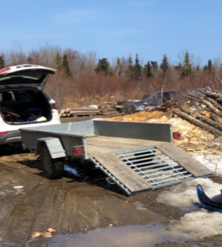 An aluminum utility trailer - with its ramp down - is attached to a white SUV. The hatch of the SUV is open and the trailer is empty;