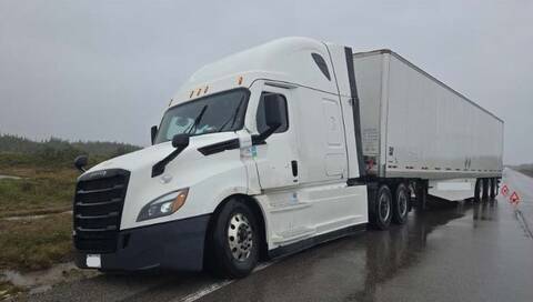 A white Freightliner commercial tractor is partially off the roadway, and its attached white trailer fully blocks one lane of the two-lane roadway.