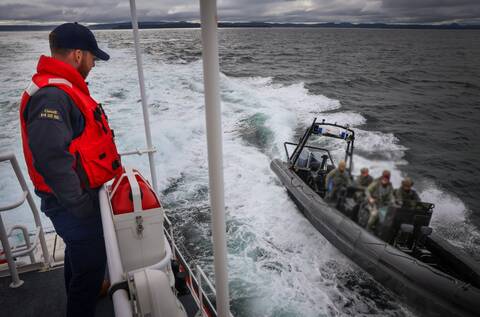 A uniformed member of the Canada Coast Guard stands on a vessel and looks on as a grey RCMP Zodiac vessel with four RCMP Emergency Response Team (ERT) officers on board pulls alongside.