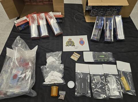 Clear plastic bags containing drugs, cell phones and other evidence are laid out on a table. Two cardboard boxes holding cartons of cigarettes are also seen in the photo. A sign reads RCMP-RNC JFO West.