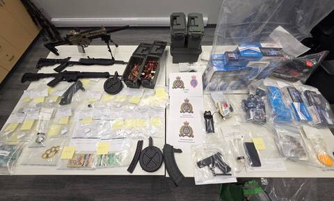 Various forms and quantities of illicit drugs, a sum of cash, contraband tobacco, multiple firearms, ammunition and a set of brass knuckles are displayed on a table. Pieces of paper, one shows the RCMP and RNC crests with “JFO West” in writing, another show the RCMP crest with “West District GIS” in writing, and another shows the RCMP Crest with “Newfoundland RCMP Rocky Harbour Detachment” in writing.