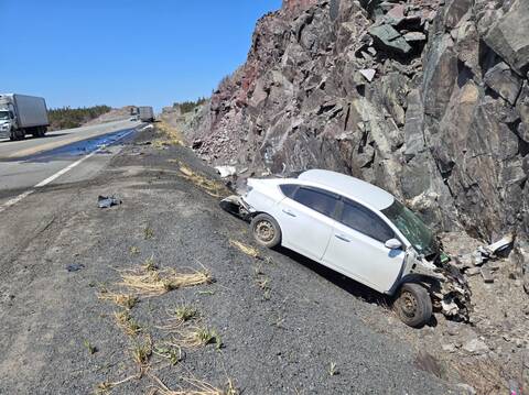 A heavily damaged white passenger vehicle is in the ditch and rests up against a rock wall. Transport trucks and other motor vehicles are visible in the background.