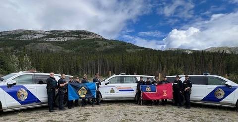 Alaska State Troopers and RCMP officers standing front of their police vehicles. RCMP officers are holding up the M Division ensign flag and the Troopers are holding their flag.