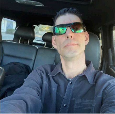 Photo of a man with a long sleeve blue collared shirt, short brown hair and sunglasses.