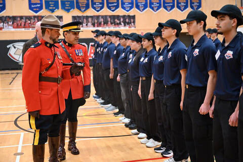 Two RCMP officers wearing red serge stand next to a row of Youth Academy students wearing baseball hats, blue collared shirts with a logo, black pants and sneakers in a school gym.