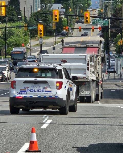 An RCMP BC Highway patrol police vehicle is driving being a large full dump truck on a busy street in Burnaby.